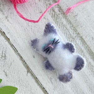 May include: A small, handmade felt cat ornament featuring a white body with light purple ears, paws, and tail. The cat has embroidered black whiskers, a pink nose, and light blue eyes, with visible white stitching along the edges of the felt.