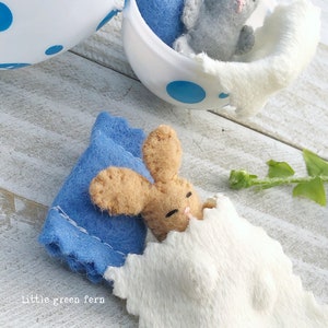 May include: Two felt bunny rabbits, one brown and one gray, are nestled in a white egg-shaped container. The brown bunny is sleeping in a blue and white blanket. The gray bunny is sitting up in a blue blanket. The egg container has blue polka dots.