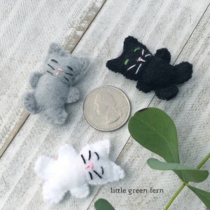 May include: Three small felt cat figures in gray, black, and white, each featuring embroidered facial details. A United States quarter coin is placed in the center for scale, measuring approximately 0.955 inches in diameter.