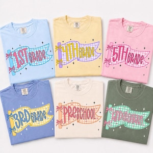 May include: Six crew neck t-shirts in light blue, yellow, pink, periwinkle, cream, and olive green. Each shirt features a graphic of a gingham-patterned flag with bows on the pole, displaying text for school grades: 1st Grade, 4th Grade, 5th Grade, 3rd Grade, Preschool, and Kindergarten.