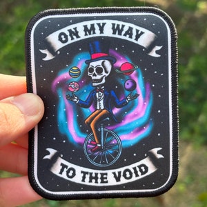May include: A black and white embroidered patch with a skeleton riding a unicycle and juggling planets. The text "On My Way To The Void" is written on the patch.