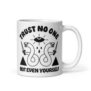 Puede incluir: Taza de cerámica blanca con un diseño gráfico negro. El diseño presenta dos serpientes con espadas, un ojo con una lágrima y el texto "TRUST NO ONE NOT EVEN YOURSELF".