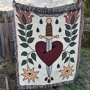 May include: Cream-colored woven throw blanket with a central design of a heart pierced by a sword. The blanket features a border of fringe and is decorated with stylized flames, leaves, stars with eyes, and raindrops. The color palette includes burgundy, brown, and green.