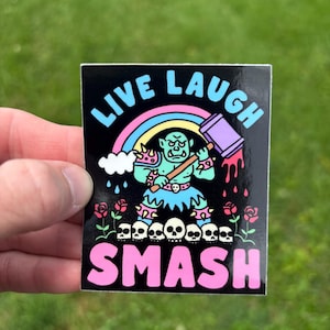 Könnte beinhalten: Schwarzer Aufkleber mit den Worten "LIVE LAUGH SMASH" in Blau und Pink. Der Aufkleber zeigt einen Cartoon-Orc-Charakter mit einem Hammer, einem Regenbogen, Totenköpfen und Rosen.