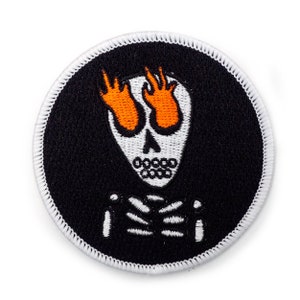 May include: A white embroidered patch with a black background. The patch features a white skeleton with orange flames for eyes. The skeleton is making the rock and roll hand gesture with both hands.