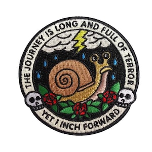 May include: Embroidered patch featuring a snail with a shell, surrounded by roses and skulls. The text reads "The journey is long and full of terror yet I inch forward".