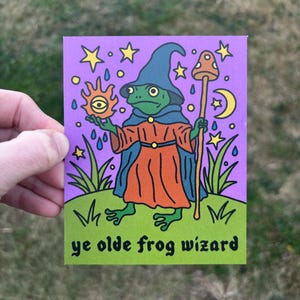 May include: A whimsical illustration of a frog wizard on a purple background. The frog wears a blue hat and robe, holding a staff and a sun with an eye. The words "ye olde frog wizard" are at the bottom, with stars and a crescent moon in the background.