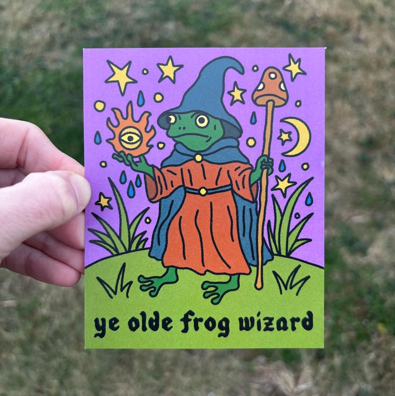 May include: A whimsical illustration of a frog wizard on a purple background. The frog wears a blue hat and robe, holding a staff and a sun with an eye. The words "ye olde frog wizard" are at the bottom, with stars and a crescent moon in the background.