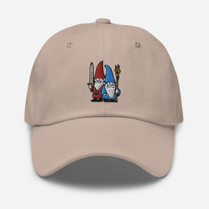 May include: A beige baseball cap featuring an embroidered design of two gnomes. One gnome is wearing a red hat and holding a sword, while the other gnome is wearing a blue hat and holding a staff.