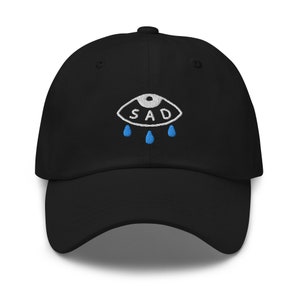 May include: Black baseball cap with an embroidered white eye with the word "SAD" inside and three blue teardrop shapes below.