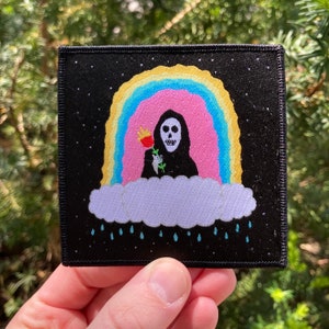May include: A black embroidered patch with a skeleton figure holding a flower, standing on a cloud with a rainbow above. Rain drops are falling from the cloud.