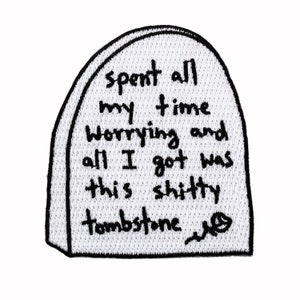May include: A white embroidered patch shaped like a tombstone with the text "Spent all my time worrying and all I got was this shitty tombstone."