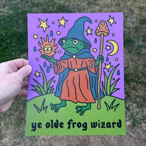 May include: A whimsical illustration of a frog wizard in a blue hat and orange robe, holding a sun with an eye and a mushroom staff. The background is purple with stars, rain, and a crescent moon. The text "ye olde frog wizard" is at the bottom.