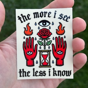 May include: A sticker with a red rose, two red hands with eyes, and an hourglass. The text reads "the more i see the less i know".