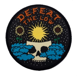 Pode incluir: Etiqueta bordada preta com uma caveira, um sol e girassóis. O texto "DEFEAT THE LOW" é escrito em um padrão circular ao redor da caveira.