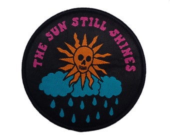 Może przedstawiać: Czarna naszywka haftowana z różowym i białym tekstem, który brzmi "The Sun Still Shines". Naszywka przedstawia kreskówkową słońce z czaszką i niebieską chmurę z kroplami deszczu.