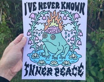 May include: A print featuring a cartoon frog with a brain on fire, surrounded by flowers and water lilies. The text reads "I'VE NEVER KNOWN INNER PEACE" in a bold, black font. The frog has spiral eyes and is in a pond.