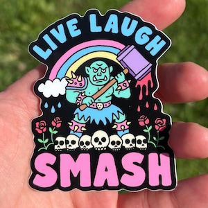 Könnte beinhalten: Ein gestanzter Aufkleber mit einem hellgrünen Ork, der einen violetten Hammer hält und über einer Reihe weißer Totenköpfe steht. Das Design enthält einen Regenbogen, rosa Rosen und den Text LIVE LAUGH SMASH in Blau und Pink auf schwarzem Grund.