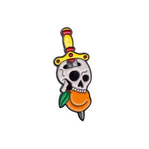 Puede incluir: Un pin de esmalte blanco y negro con una calavera con una daga atravesando su cabeza y un melocotón en su boca. La daga tiene un mango amarillo y detalles rojos. El melocotón es naranja con una hoja verde.