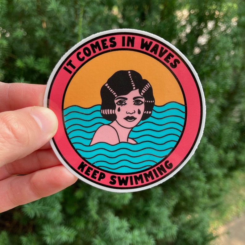 Op de afbeelding: Een ronde sticker in roze en wit met de tekst "It comes in waves keep swimming" in zwart-witte letters. De sticker heeft een cartoonillustratie van een vrouw met zwart haar en een blauw-wit golfpatroon.