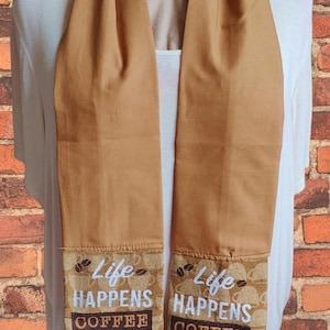 May include: A tan coffee-themed scarf with the text "Life Happens Coffee Helps" and a coffee cup design. The scarf is displayed against a brick wall background.