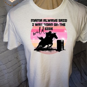 Pode incluir: Camiseta branca com um gráfico de pôr do sol rosa, laranja e amarelo. O gráfico apresenta uma silhueta de uma pessoa montando um cavalo e um barril. O texto "Mama Always Said I Was Born On The Wild Side" está impresso em preto acima do gráfico.