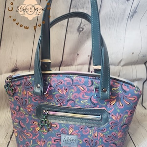 May include: A teal blue handbag with a paisley print and black leather accents. The bag has a zipper closure and a top handle. The bag has a small pocket on the front with the text "S.Kay Designs" in pink.