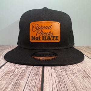 May include: Black snapback hat with a flat brim. The hat features a rectangular, tan leather patch with the words "Spread Cheeks Not HATE" in a script font. The hat is sitting on a wooden surface.