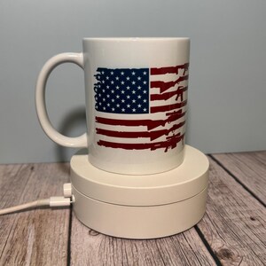 May include: White ceramic mug featuring a distressed American flag design with red and blue accents. The flag design incorporates the stars and stripes, along with silhouettes of rifles. The mug has a curved handle and is displayed on a white rotating stand.