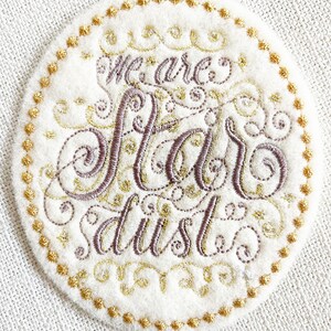 May include: A white circular patch with a gold beaded border. The text "We are star dust" is embroidered in gold and brown thread.