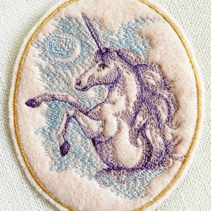 May include: Embroidered patch featuring a purple unicorn with a gold horn, standing on a cloud with a crescent moon in the background. The patch is surrounded by a gold border.