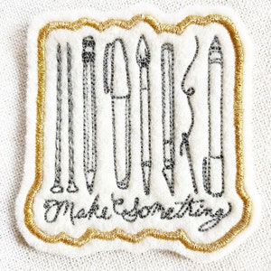May include: A gold embroidered patch with a white background featuring a collection of drawing tools, including a pencil, pen, and paintbrush. The text "Make Something" is embroidered in black below the tools.