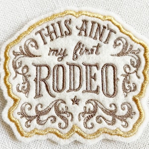 May include: A white felt patch with the embroidered phrase "THIS AIN'T my first RODEO" in brown script. The patch has a gold scalloped border and decorative flourishes. A small star is also embroidered on the patch.