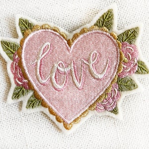 May include: A pink felt heart-shaped patch with gold embroidery around the edges. The word "love" is embroidered in white on the heart. The heart is surrounded by green and pink felt flowers.