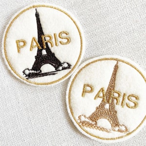 May include: Two round, white felt patches with gold embroidery. Each patch features a black and gold silhouette of the Eiffel Tower and the word "PARIS" in gold embroidery.