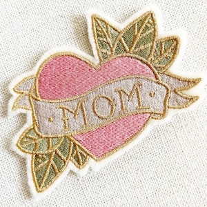 May include: A pink heart with a gold outline and a banner that says "MOM" in gold lettering. The heart is surrounded by green leaves with gold outlines.