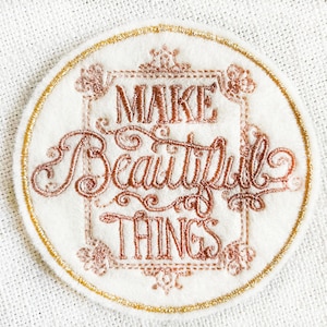 May include: A white round embroidered patch with a gold border. The patch features the text "MAKE BEAUTIFUL THINGS" in a brown cursive font.