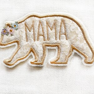 May include: A white lace bear applique with gold embroidery. The bear has a floral crown and the word "MAMA" embroidered on its back.