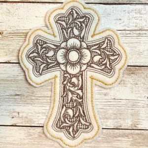May include: A white cross with a gold outline and a brown floral design. The cross is embroidered with a detailed floral pattern and a large flower in the center.