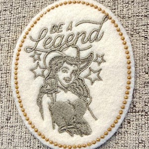 May include: Oval-shaped patch with a white background, featuring an embroidered design of a woman in a cowboy hat, stars, and the words "BE A Legend". The patch is bordered with gold beads.