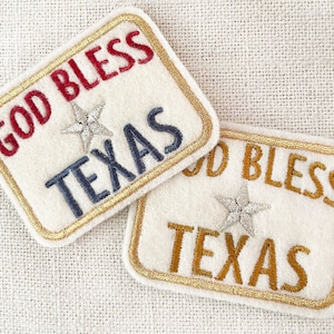 May include: Two white felt patches with gold trim and embroidered text. The top patch reads "God Bless Texas" in red and blue lettering with a silver star. The bottom patch reads "God Bless Texas" in gold lettering with a silver star.