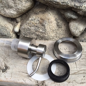 Peut inclure: Une pompe de distributeur de savon en acier inoxydable avec un tube en plastique transparent, un anneau métallique, un joint blanc et un joint noir. Le distributeur est conçu pour s'adapter à un bocal. Les articles sont présentés sur une surface en bois.