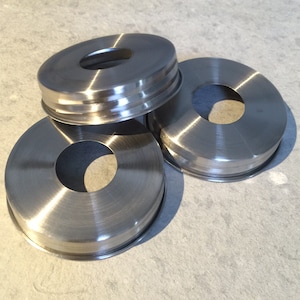 May include: Three silver metal lids with a hole in the center. The lids are stacked on top of each other.