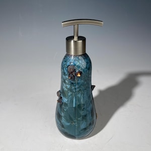 May include: A decorative soap dispenser with a blue and teal crystalline glaze. The dispenser has a brushed metal pump and is adorned with dark brown, textured embellishments. The bottle's shape is elongated, with a rounded base.