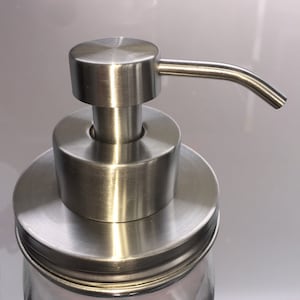 Peut inclure: Un distributeur de savon en acier inoxydable avec un bec verseur incurvé. La pompe est fixée à un bocal en verre transparent. Le distributeur a une finition en métal brossé et un dessus rond.