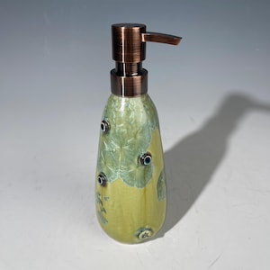 May include: A ceramic soap dispenser with a copper-colored pump. The bottle is a teardrop shape, with a light green and yellow glaze. It has small, dark blue accents and a textured pattern.