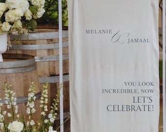 May include: A cream-colored wedding welcome sign with the names "Melanie & Jamaal" and the text "You look incredible, now let's celebrate!" The date "10.19.2025" is also displayed. The sign is on a white metal frame.