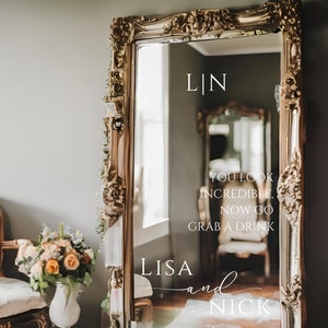 May include: A gold-framed mirror with a white vinyl decal that reads "LIN", "YOU LOOK INCREDIBLE, NOW GO GRAB A DRINK", "LISA and NICK", and "05.25.2025". The mirror is leaning against a wall with a bouquet of flowers in a vase in the foreground.