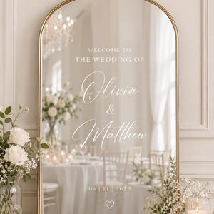 Puede incluir: Espejo dorado con marco arqueado, con el texto «Welcome to the Wedding of Olivia & Matthew» y la fecha 06|11|2025. El espejo refleja una recepción de boda con flores y velas.