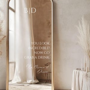 Puede incluir: Un gran espejo dorado adornado con un marco dorado y una parte superior arqueada. El espejo refleja una habitación con una cortina blanca y el texto "YOU LOOK INCREDIBLE! NOW GO GRAB A DRINK." El texto está en una fuente delicada.